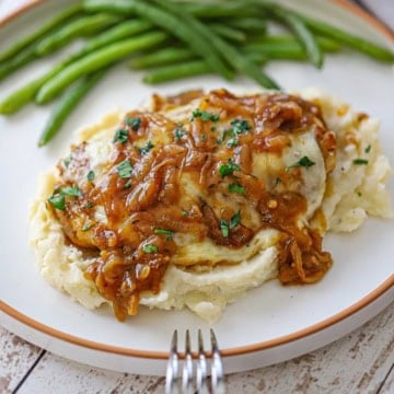 A close-up view of a French onion-smothered chicken on a bed of mashed potatoes with steamed green beans in the background, all on a white dinner plate.