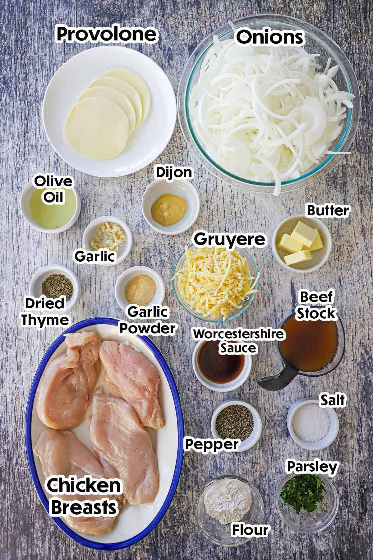 An arrangement of ingredients for French onion mothered chicken on a grey wooden background including chicken breasts, onions, beef stock, garlic, flour, oil, cheese, and seasonings.