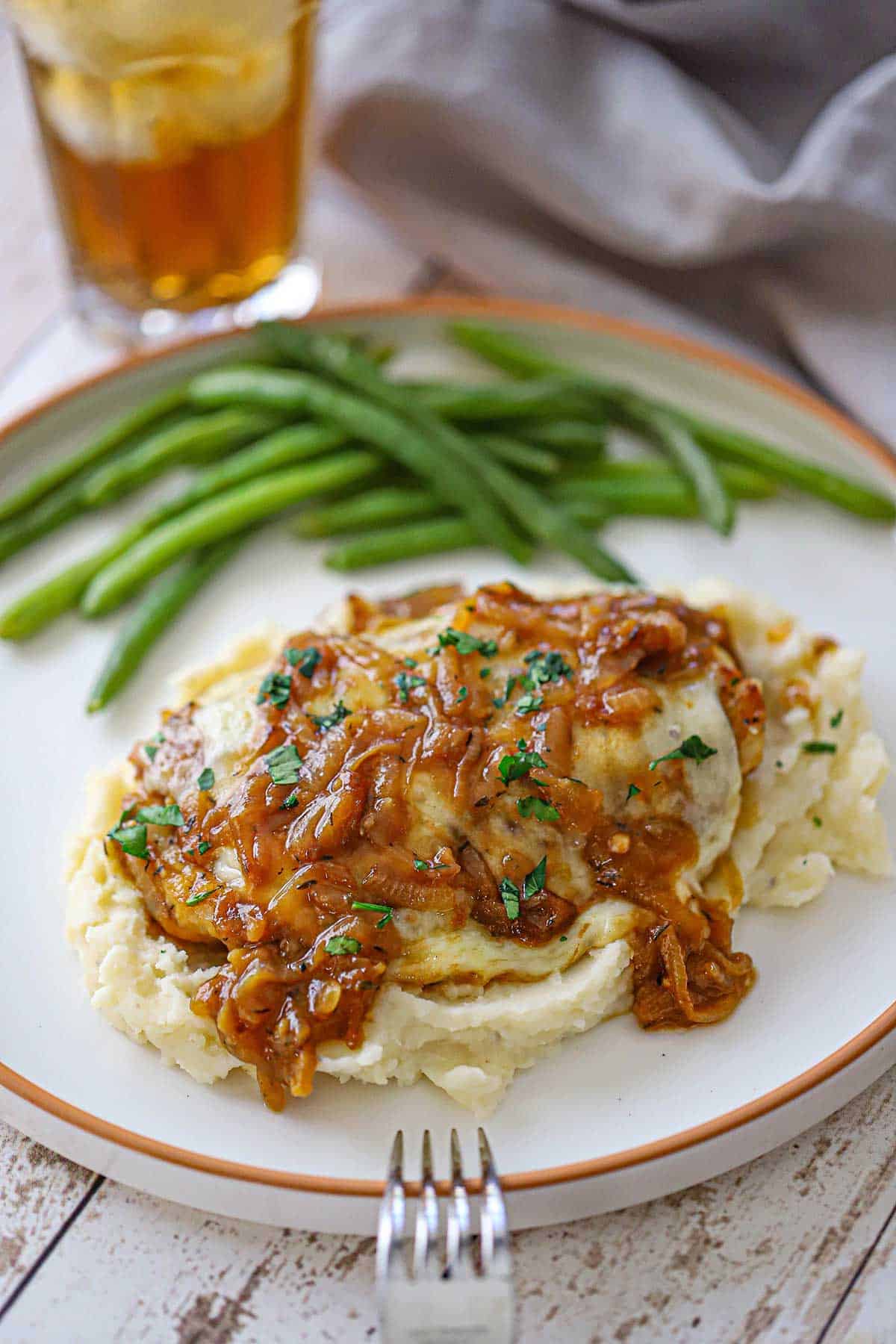 A French onion-smothered chicken on a bed of mashed potatoes with steamed green beans in the background, all on a white dinner plate.