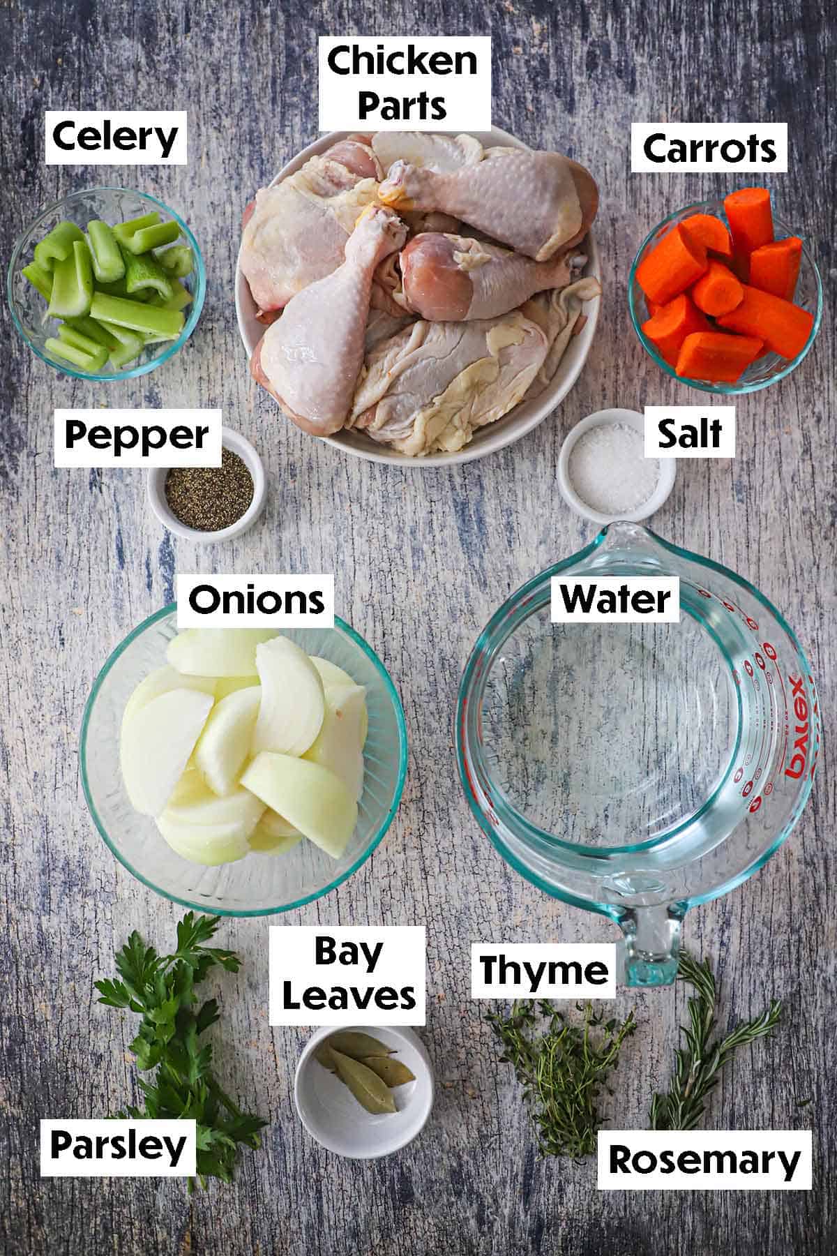 An arrangement of ingredients for Homemade Chicken Broth on a grey wooden background including raw chicken pieces, celery, carrots, onions, water, fresh herbs, and seasonings.