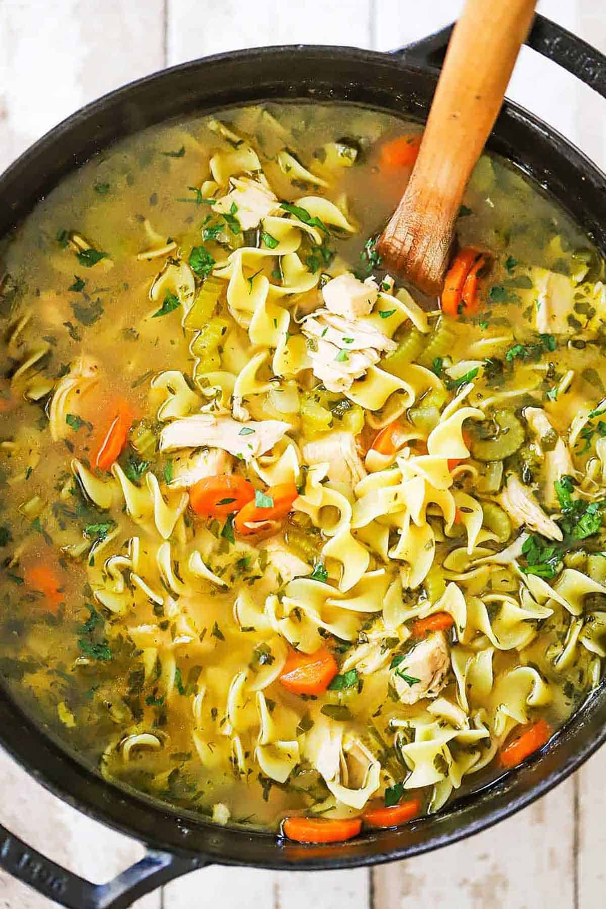 An overhead view of a black pot filled with homemade chicken noodle soup with a wooden spoon inserted into the soup.