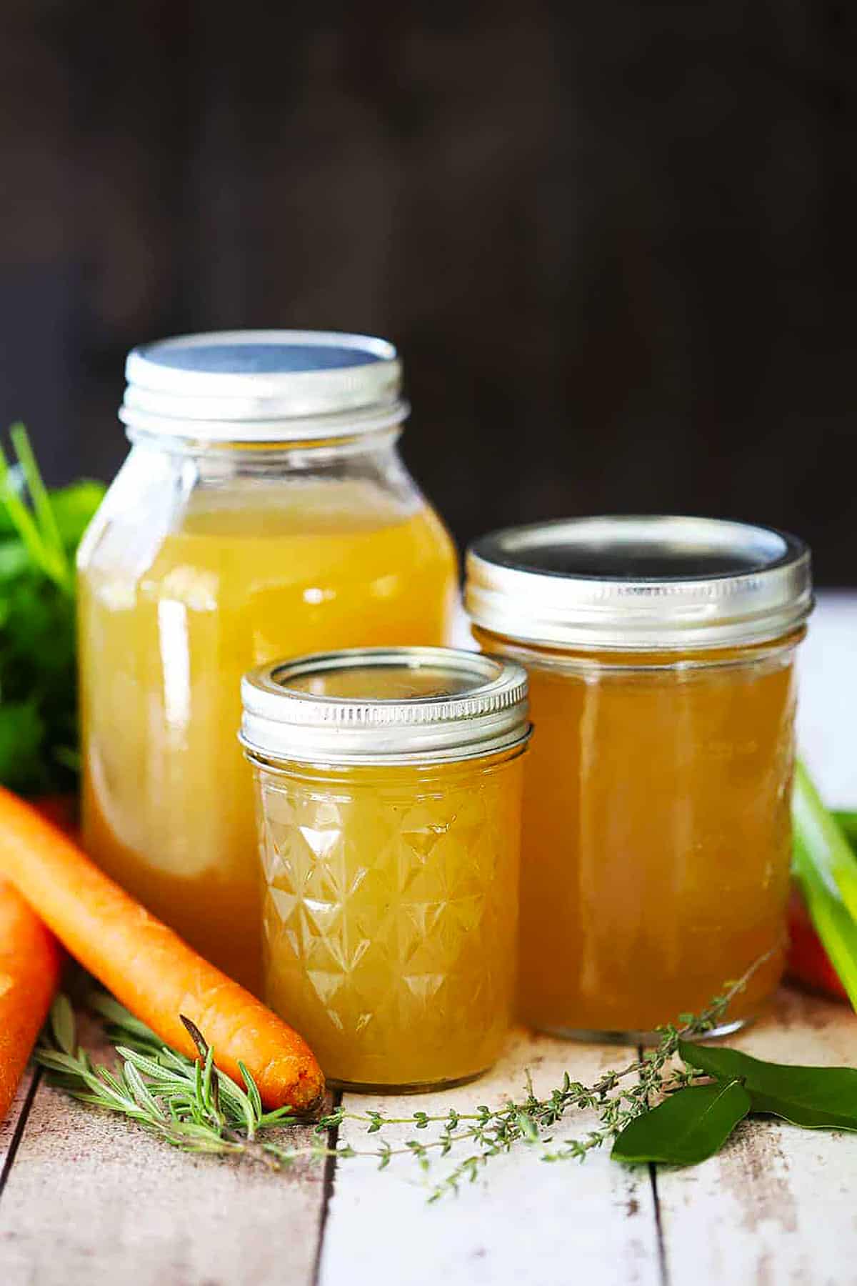 A straight-on view of three varying sizes of glass jars filled with homemade chicken broth along with carrots, celery, and herbs along the bottoms of the jars.