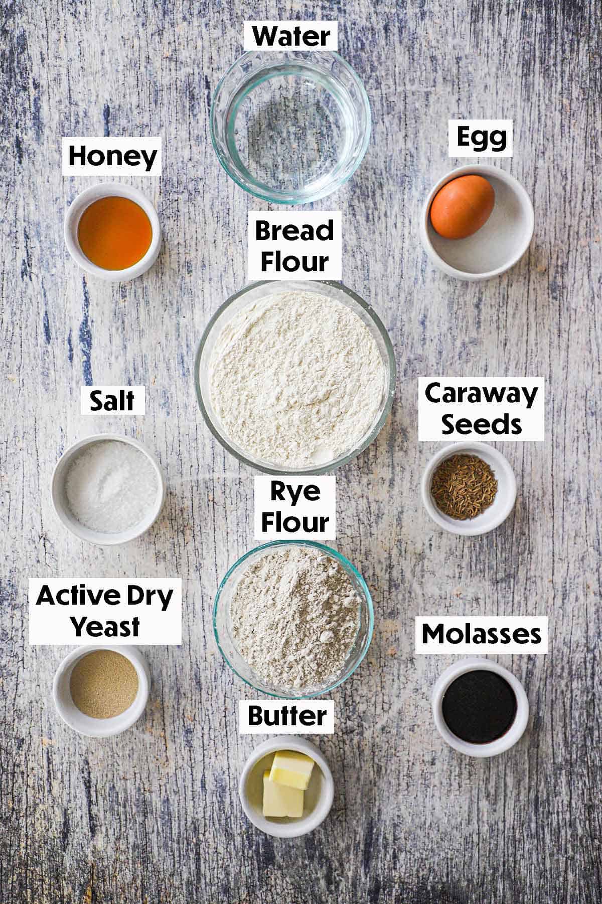 An arrangement of ingredient for deli-style rye bread on a grey wooden background including rye flour, bread flour, water, honey, an egg, yeast, molasses, butter, salt, and caraway seeds.