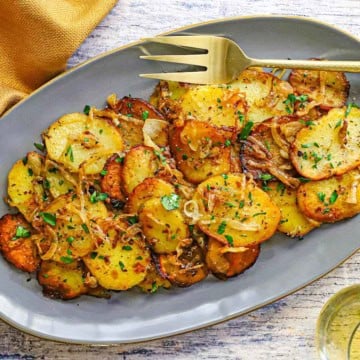 An overhead view of a grey, oval serving platter filled with a pile of freshly prepared Authentic Lyonnaise Potatoes with a large gold serving fork sitting on the edge of the platter.