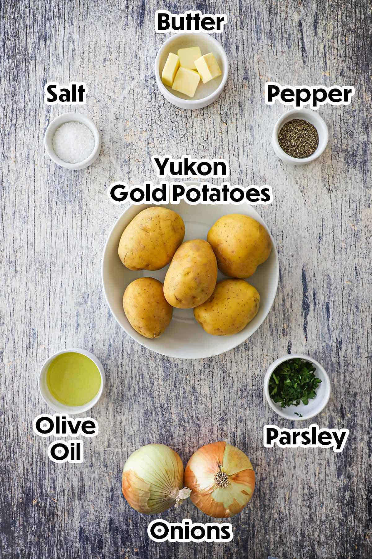An arrangement of ingredients for Authentic Lyonnaise Potatoes on a grey wooden background including Yukon gold potatoes, onions, butter, olive oil, salt, pepper, and parsley.