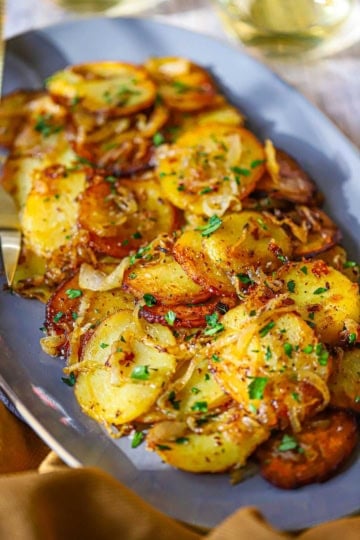 A close-up view of an oval grey serving platter filled with a pile of Authentic Lyonnaise Potatoes garnished with chopped Italian parsley.