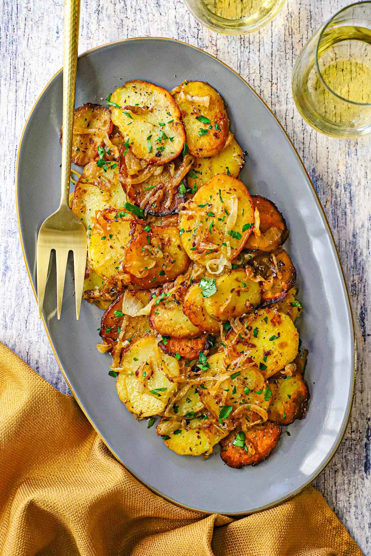An overhead view of a grey, oval serving platter filled with a pile of Authentic Lyonnaise Potatoes with a gold serving fork nearby and two stemless glasses of white wine next to the dish.