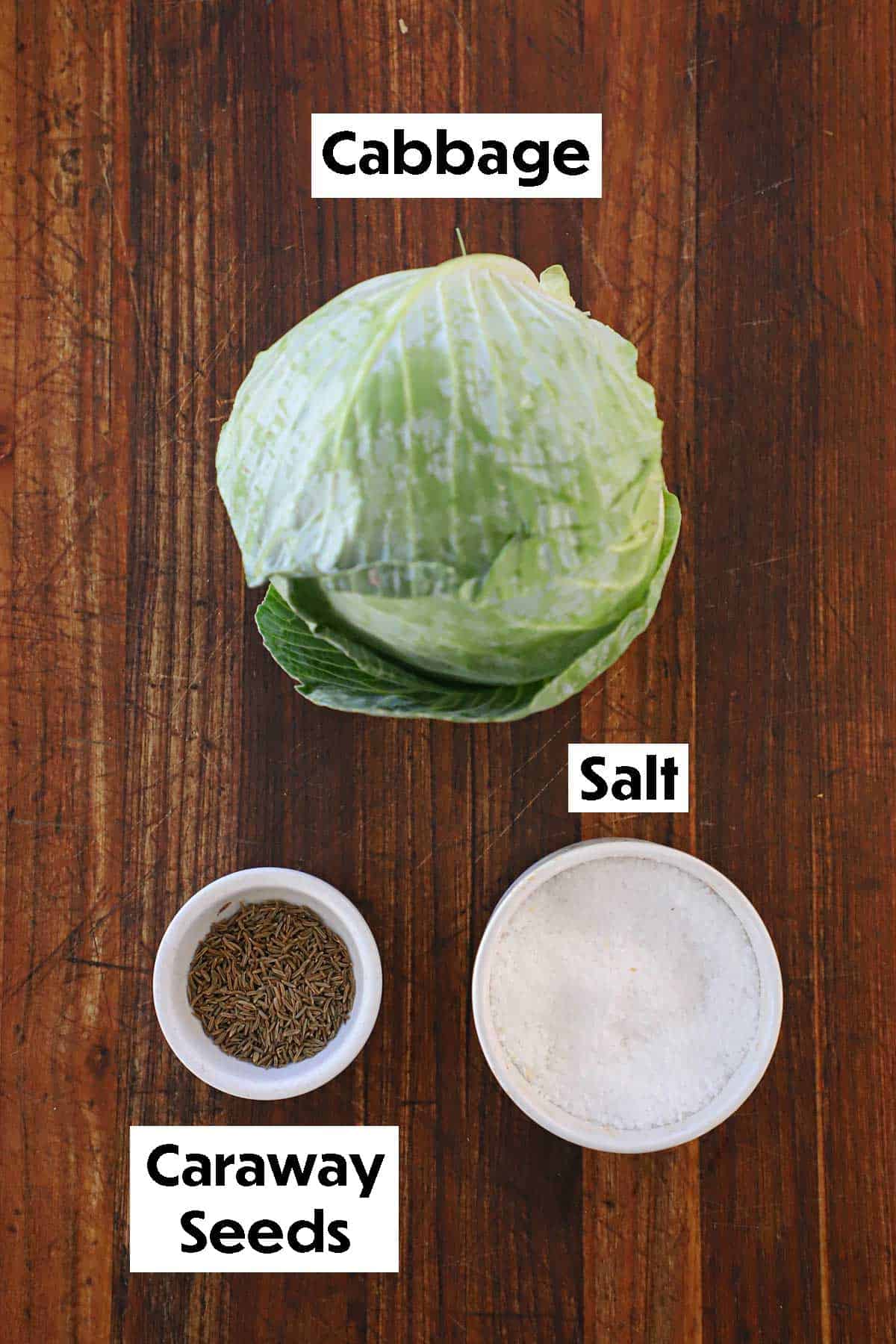 An arrangement of ingredients for homemade sauerkraut on a wooden board including head of green cabbage, caraway seeds, and salt.