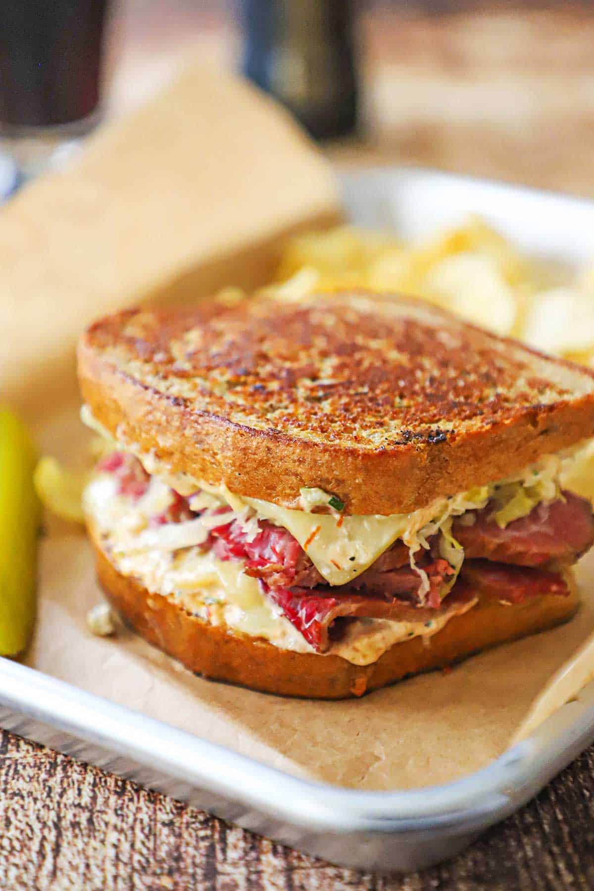 A close-up view of a "From Scratch" Reuben Sandwich resting on a brown paper in a small metal sheet pan with a pickle spear and kettle chips nearby.