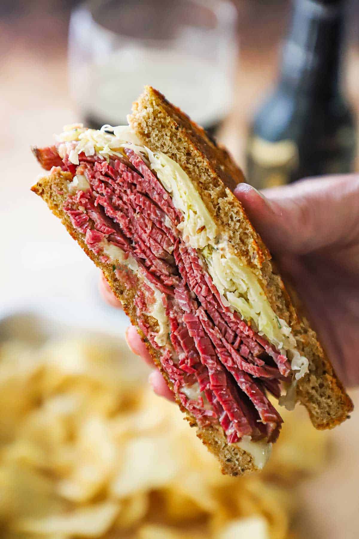 A person holding half of a "From Scratch" reuben sandwich revealing the stacks of corned beef, and the sauerkraut, melty cheese, surrounded by the toasty slices of rye bread.