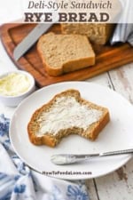 A slice of deli-style rye bead with a large bite out of it and slathered with softened butter on a white plate next to the loaf of bread on a cutting board.