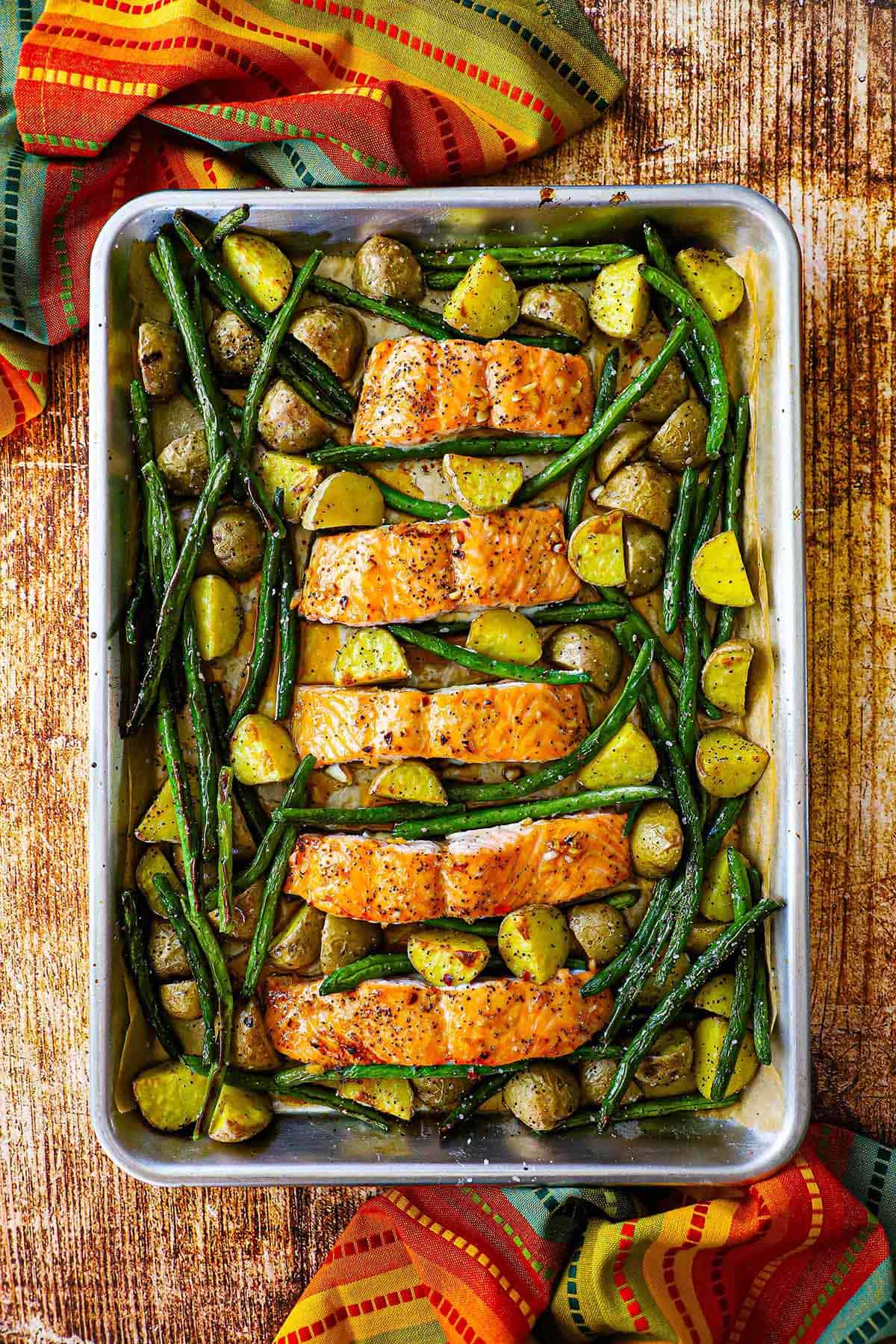 An overhead view of a large serving of sheet pan honey garlic salmon and roasted veggies in a large silver pan including roasted green beans and baby potatoes.