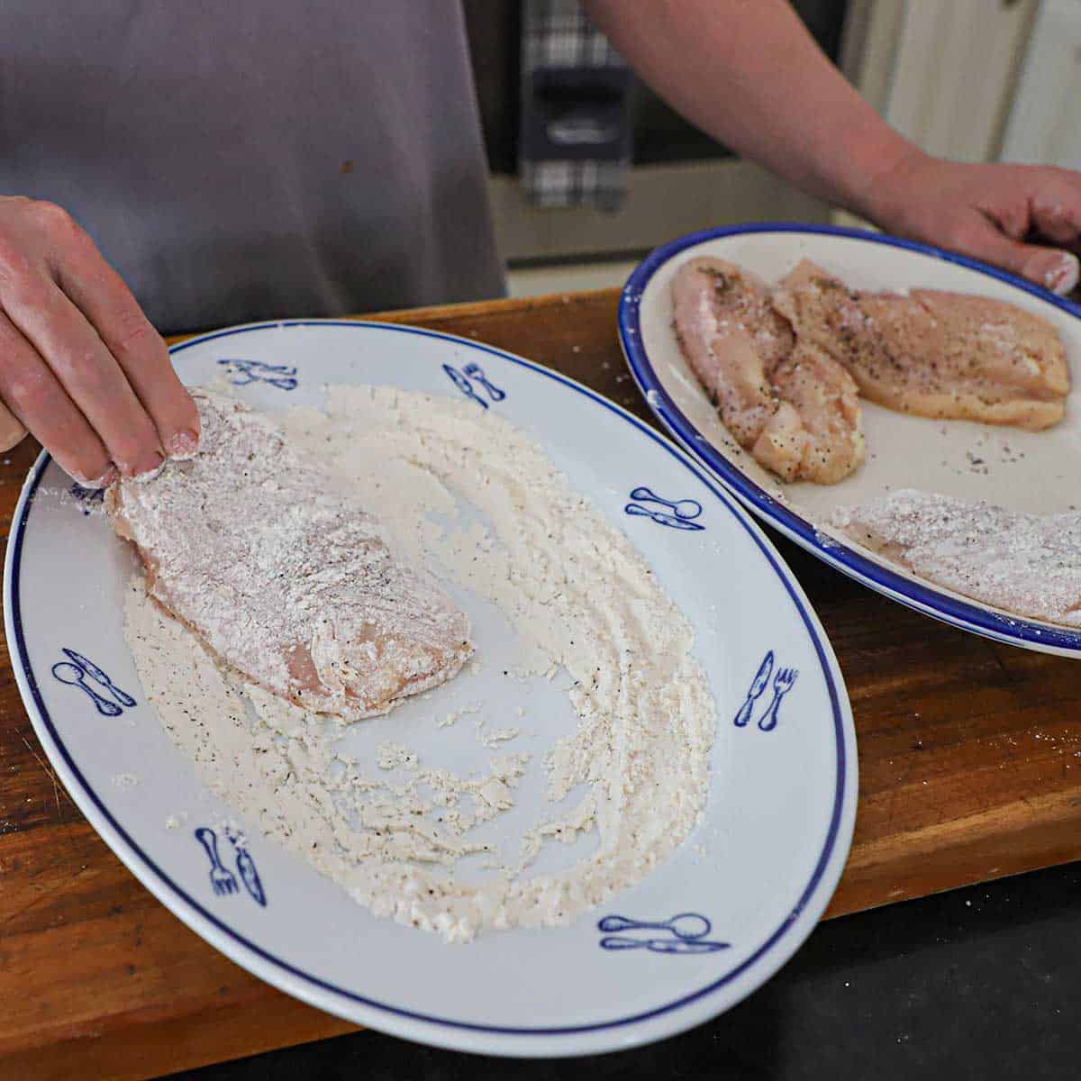 A person dredging a thin chicken breast through seasoned flour on a large platter on a wooden cutting board.