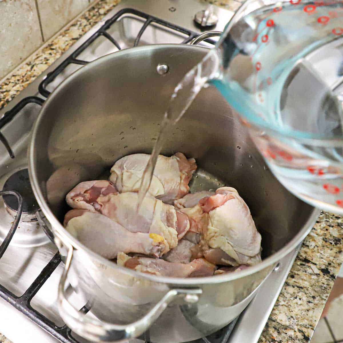 A person pouring water from a large glass measuring cup into a large stock pot filled with raw chicken pieces over a gas stove.