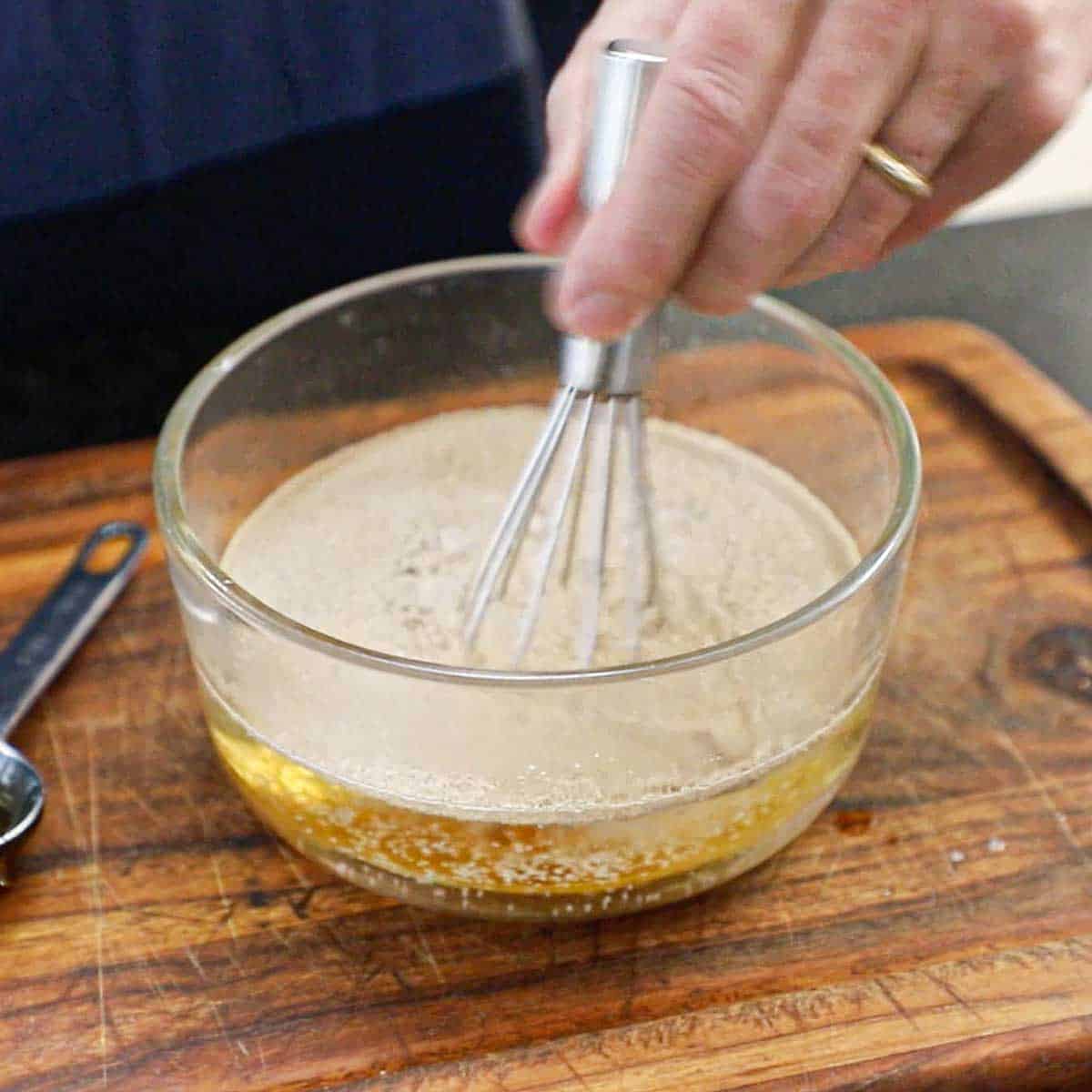 A person using a small whisk to combine warm water with honey and active dry yeast on a wooden cutting board.