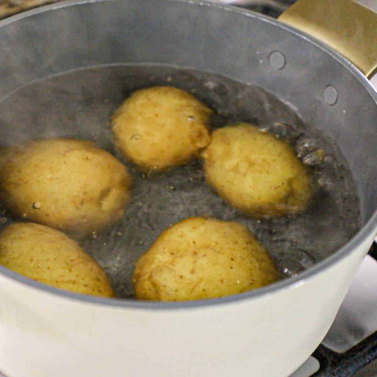 Five whole, unpeeled gold potatoes in a pot of boiling water on a gas stove.