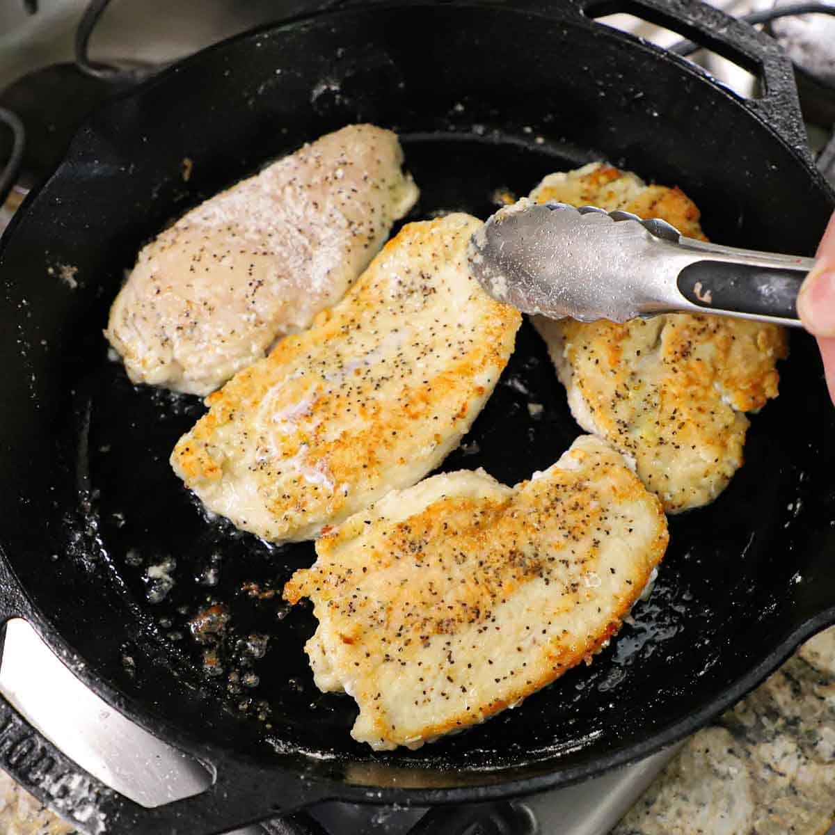 A person searing lightly breaded chicken breasts in butter and olive oil n a large cast iron skillet on a gas stove.