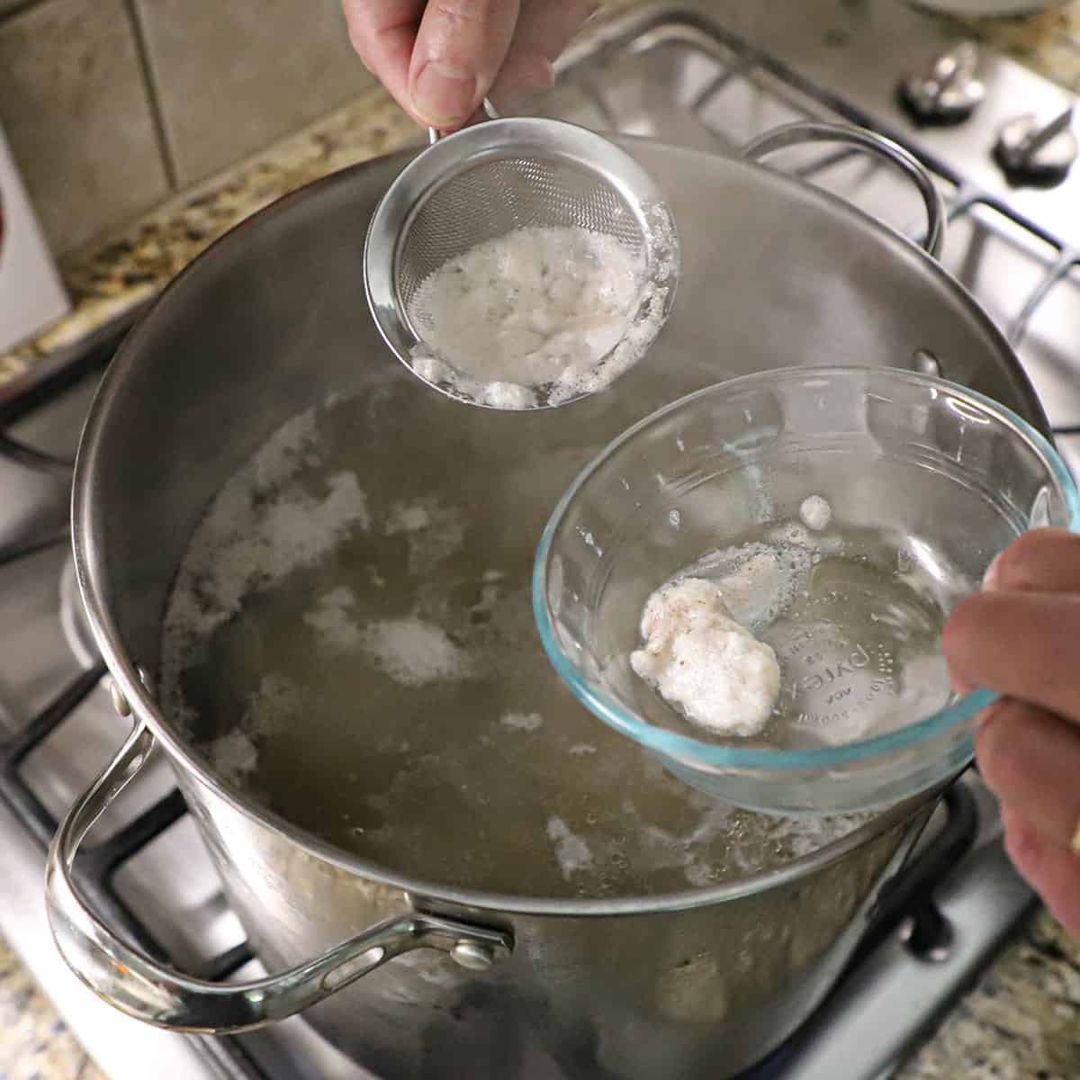 A person using a small sieve to scoop impurities that are floating on the surface of water and chicken that are being simmered in a pot on a gas stove.