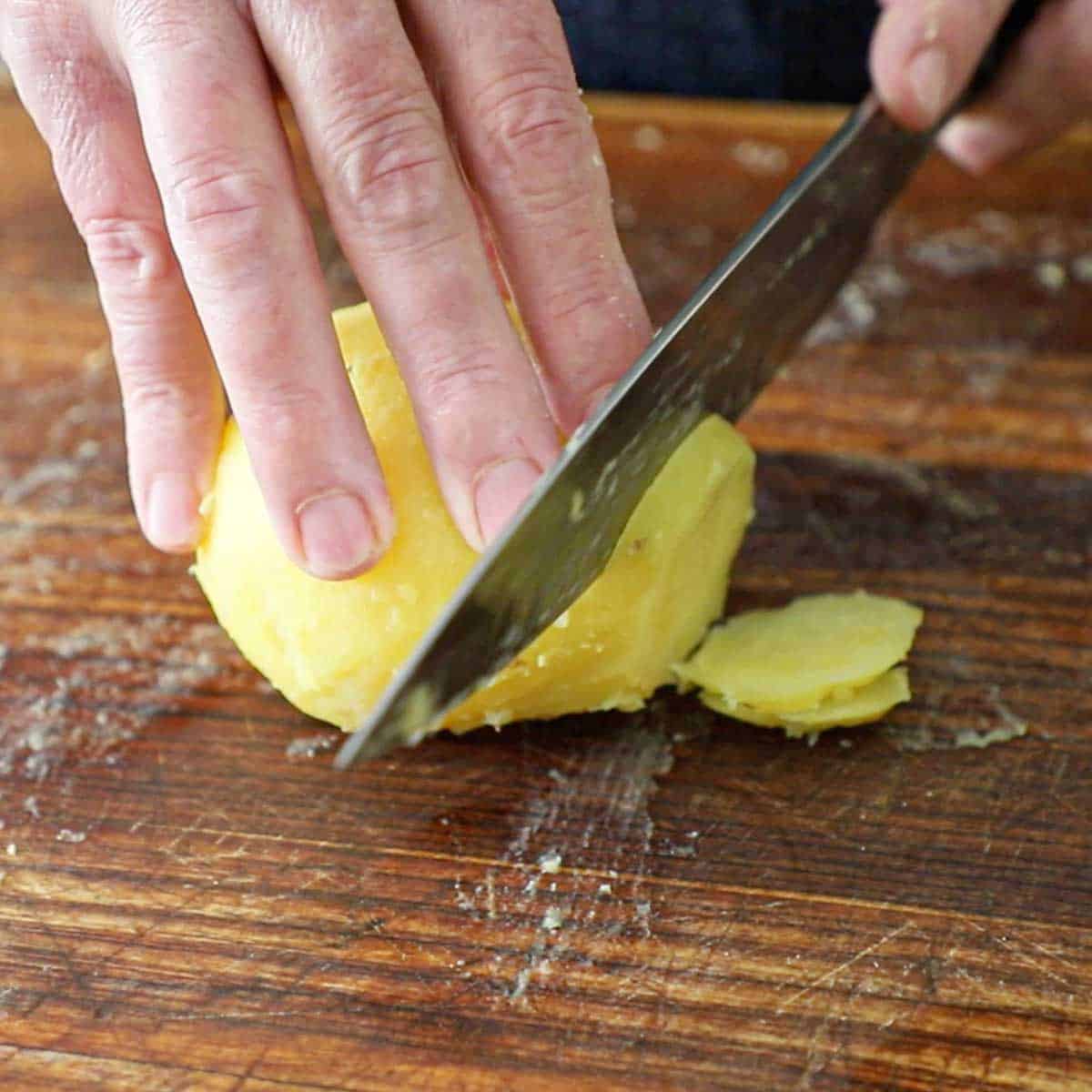 A person using a chef's knife to cut a par-boiled gold potato into slices on a large wooden cutting board.