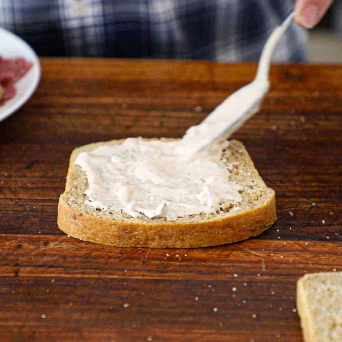 A person using a spoon to spread homemade Russian dressing over the top of a slice of rye bread on a wooden cutting board.