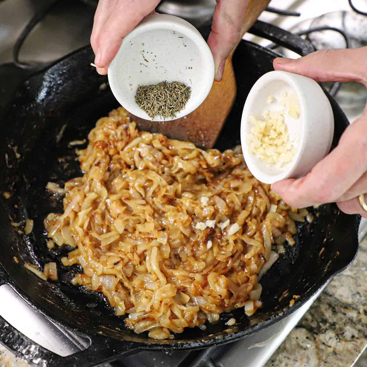 A person dumping dried thyme from a small bowl in one hand and minced garlic from a small bowl in his other hand, both going into a skillet filled with deeply caramelized onions.