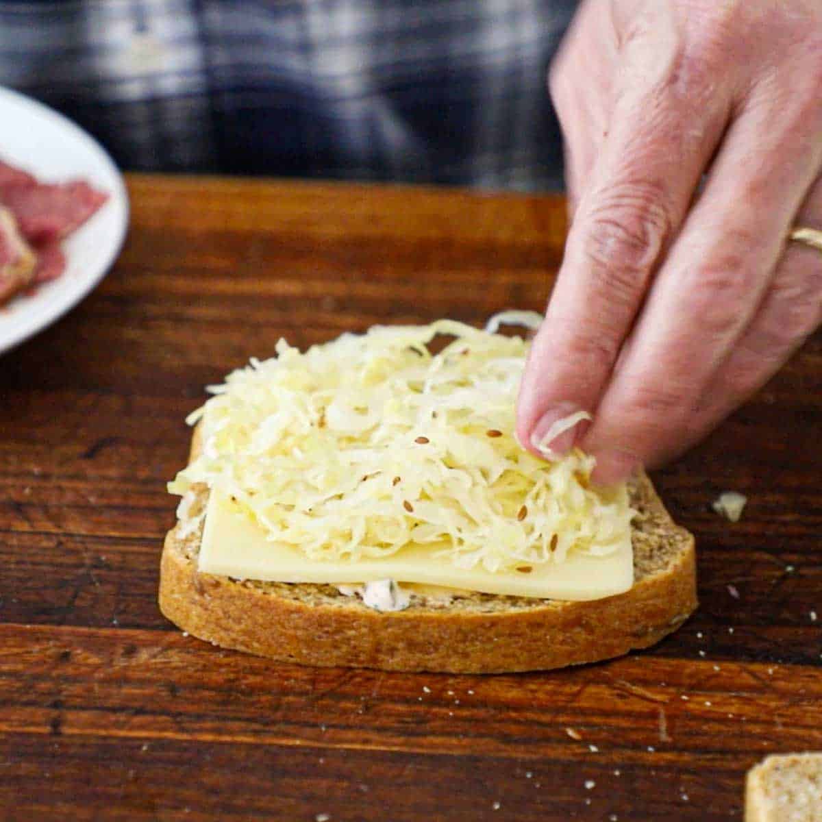 A person placing homemade sauerkraut over the top of a thick slice of Swiss cheese that is resting on a slice of rye bread and a smear of Russian dressing.