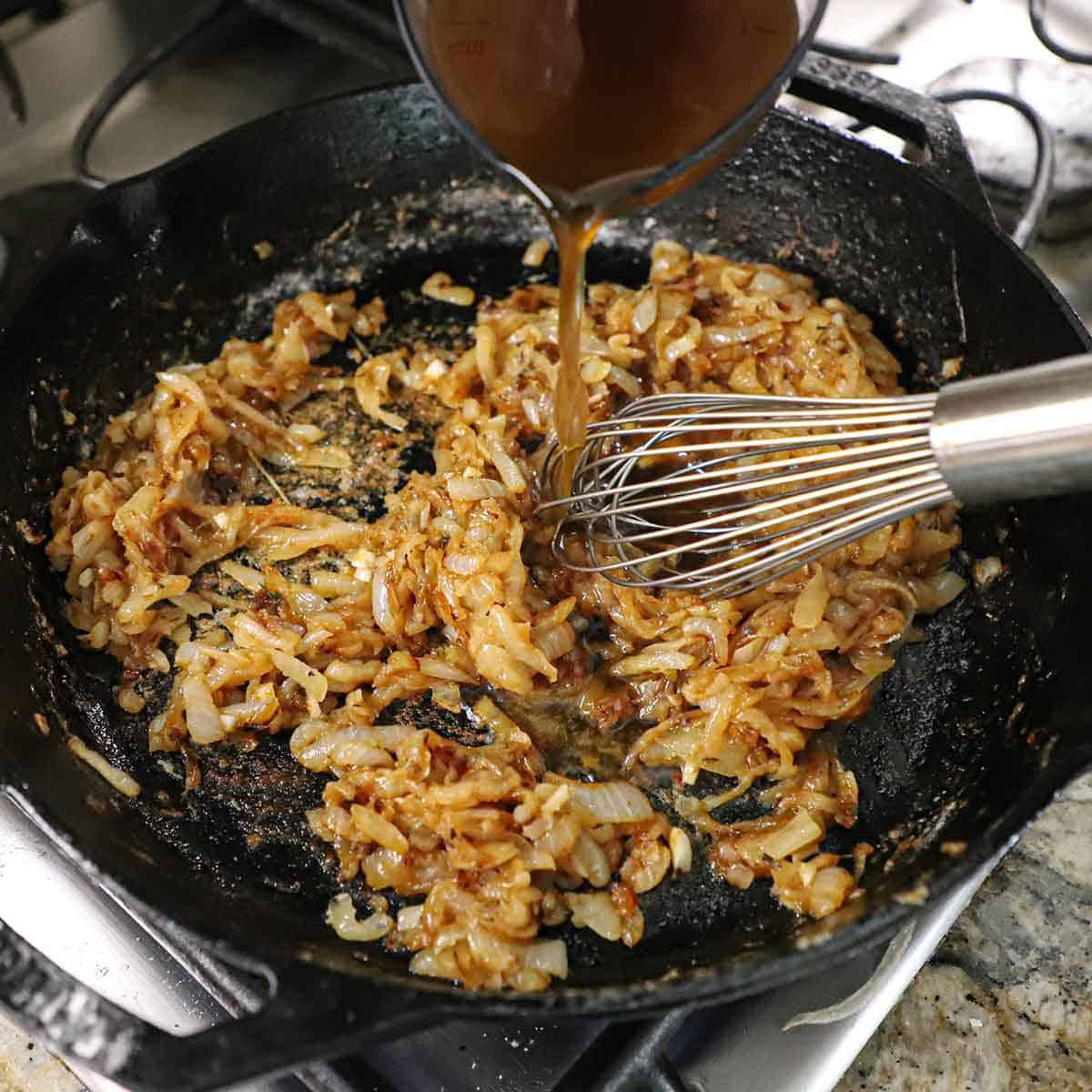 A person pouring beef stock from a clear measuring cup into a large cast-iron skillet filled with caramelized onions.
