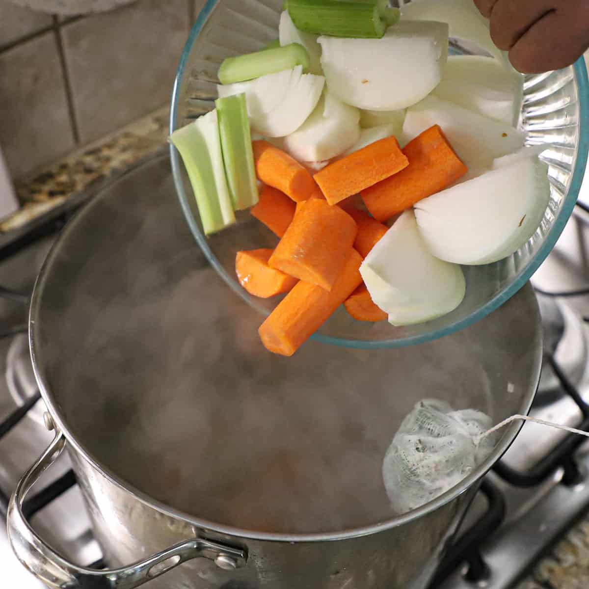 A person transferring roughly cut carrots, celery, and onion from a glass bowl into a large pot filled with simmering water, chicken pieces, and a bouqet garni.