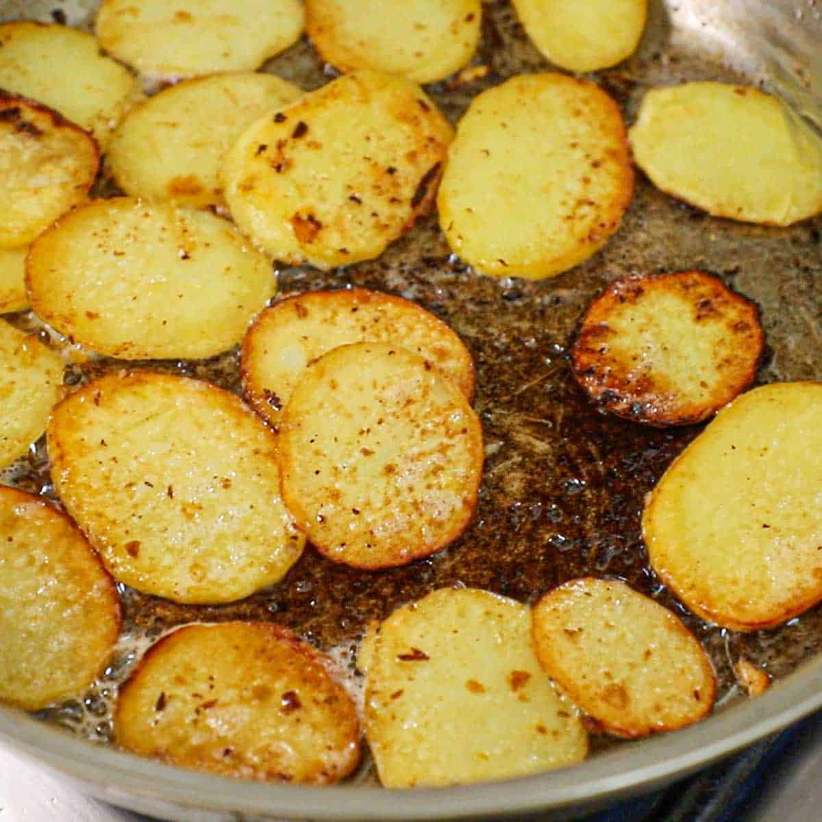 A close-up view of slices gold potatoes that are being crisped and browned in butter in a large skillet on a gas stove.