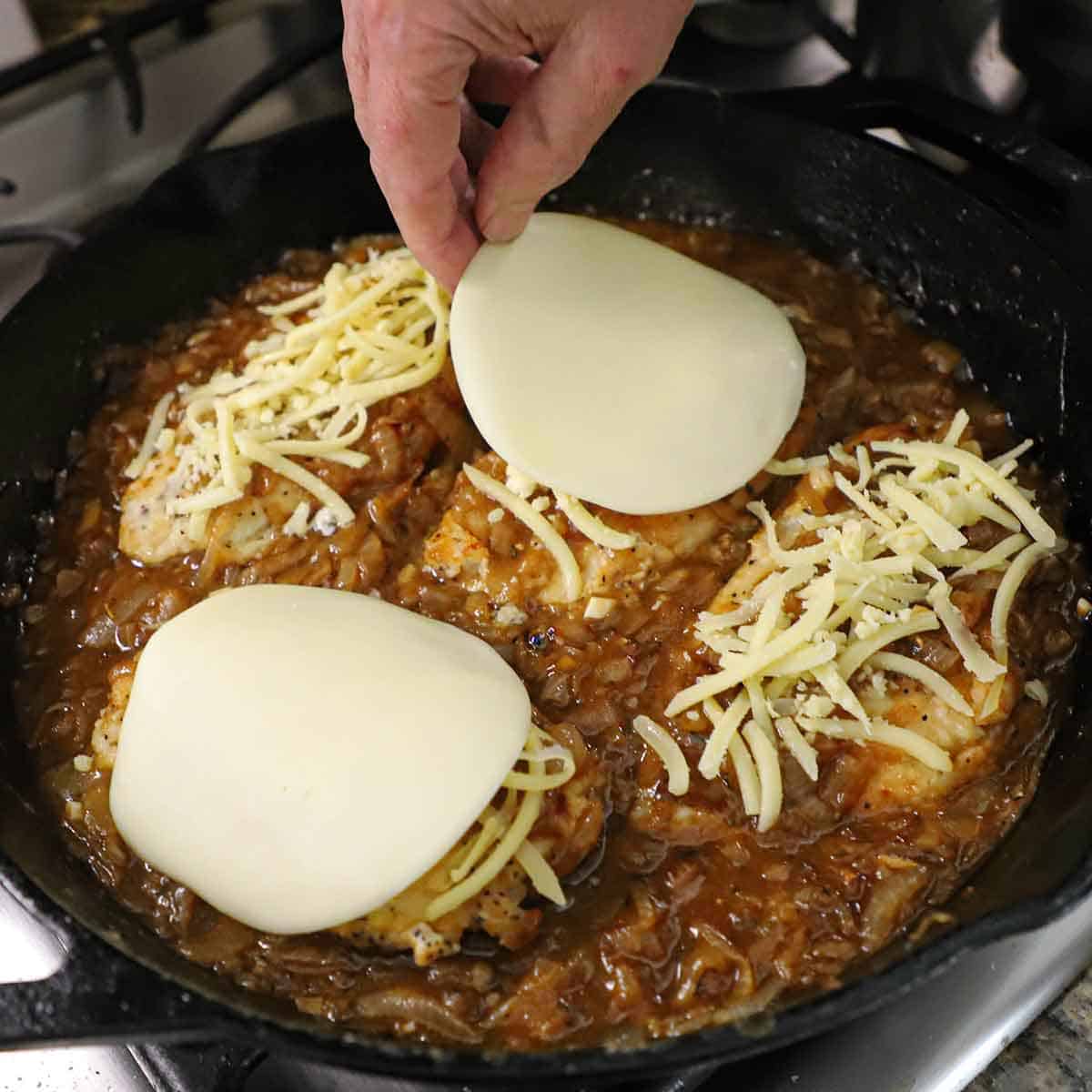 A person placing a round slice of Provolone cheese over a small mound of grated Gruyere cheese that is piled on a seared chicken breast that is nestled in a French onion sauce all in a large cast-iron skillet.