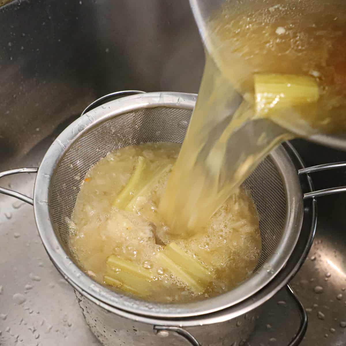 A person pouring homemade chicken broth from a large pot into a smaller pot that is lined with a fine-meshed sieve.