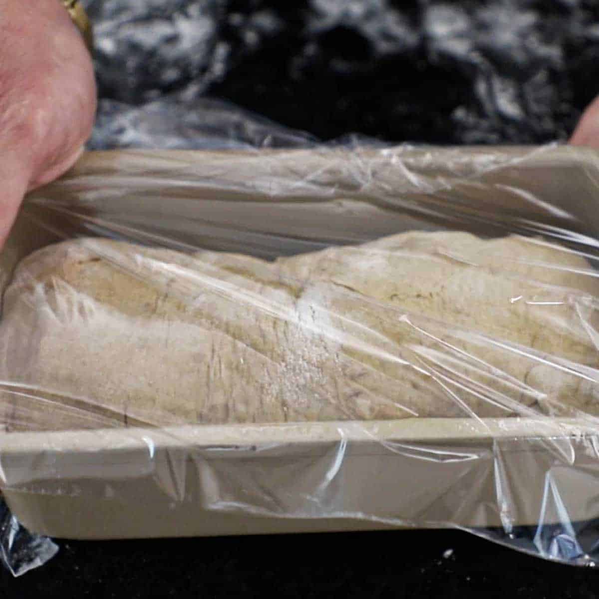 a person placing a rectangular piece of plastic wrap over a loaf pan that if filled with rye bread dough that has been shaped into a loaf.