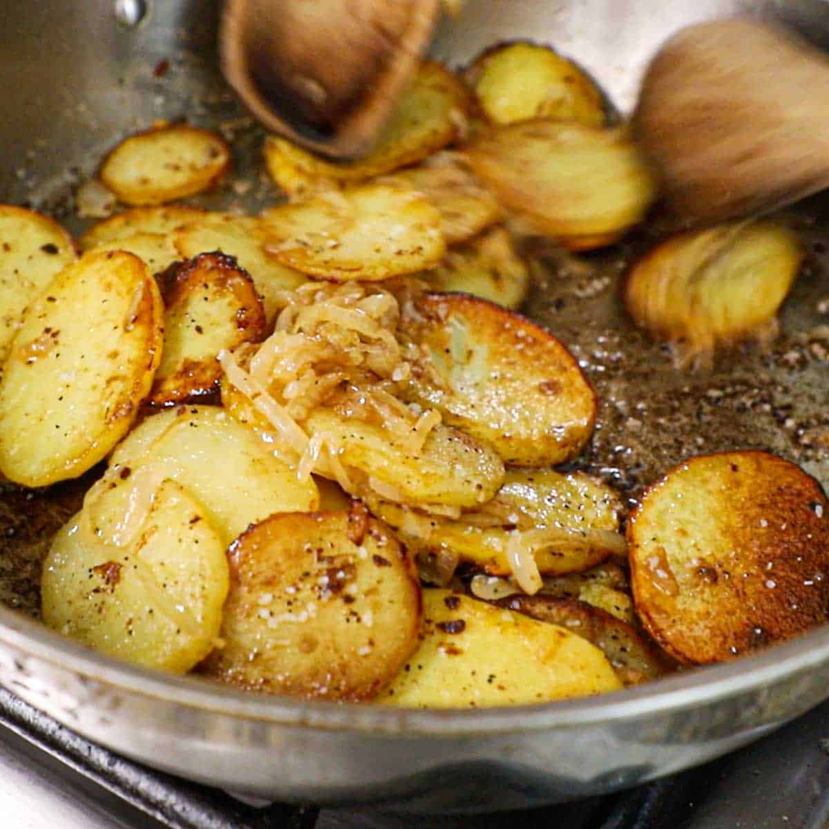 A close-up view of crispy, browned gold potato slices in a skillet that are being tossed with lightly caramelized onions.