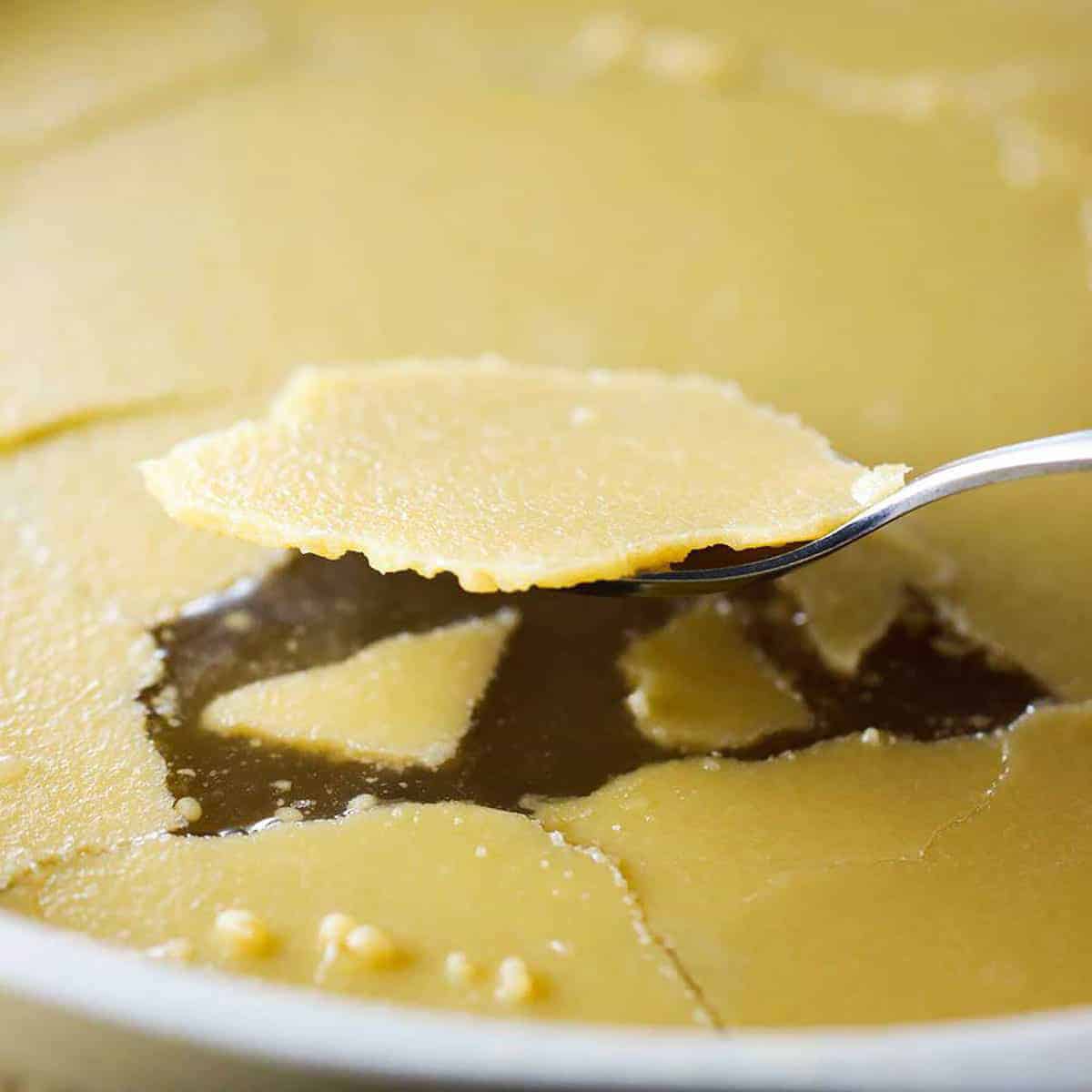 A person using a spoon to scraped up congealed chicken fat from the surface of chilled homemade chicken broth in a chilled stock pot.