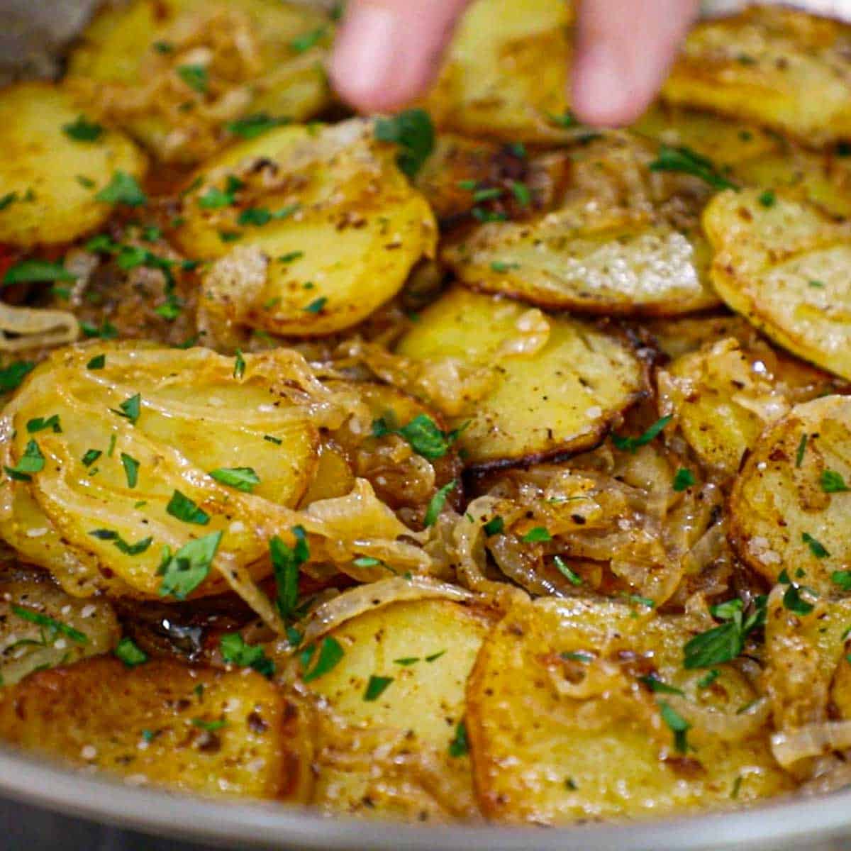 A person sprinkling chopped Italian parsley over the top of freshly prepared Authentic Lyonnaise Potatoes in a large stainless steel skillet.
