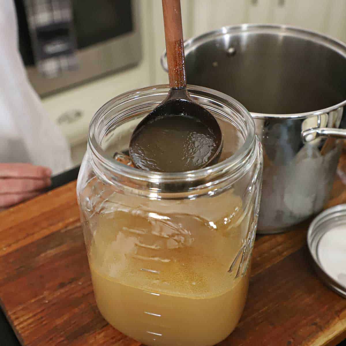 A person using a large wooden ladle to transfer homemade shellfish stock from a stock pot into an overside glass Mason jar.