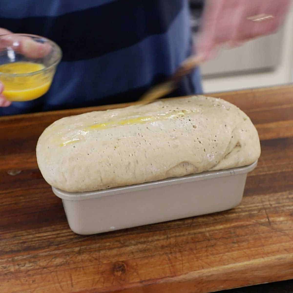 A person brushing an egg wash over a freshly risen deli-style rye bread dough in a loaf pan on a wooden cutting board.