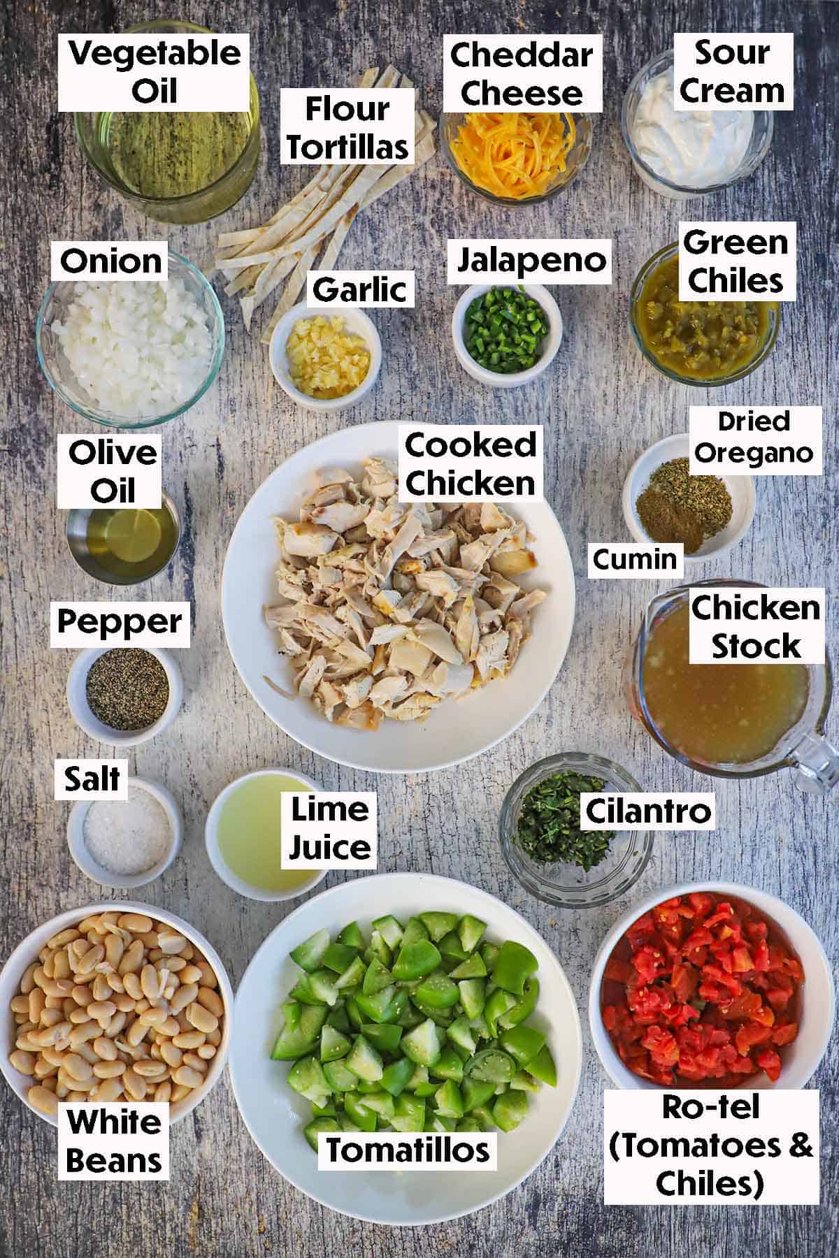 An arrangement of ingredients for white chili with tomatillos and chicken on a grey wooden background including cooked cubed chicken, chicken stock, tomatillos, diced tomatoes, white beans, green chiles, garlic, onion, oil, flour tortillas, cheese, and seasonings.
