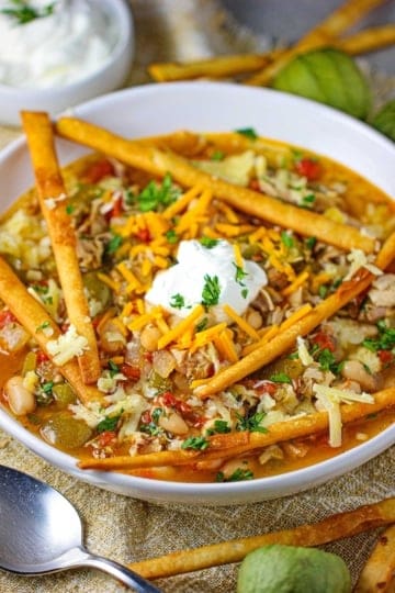 An overhead view of a white bowl filled with a serving of white chili with tomatillos and chicken topped with sour cream, chopped cilantro, and fried flour tortilla strips.