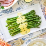An overhead view of a white rectangle platter filled with a dish of Asparagus with Béarnaise sauce and a large gold serving fork nearby.