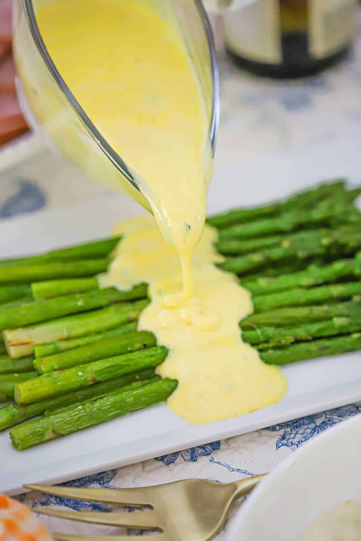 A person pouring blender Béarnaise sauce from a glass gravy boat of a pile of pan-seared asparagus on a white platter.