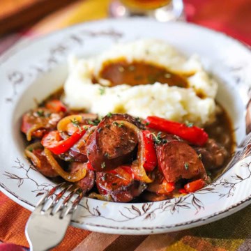 A close-up view of a vintage soup bowl filled with beer-braised kielbasa and peppers next to a pile of mashed potatoes with gravy on top.