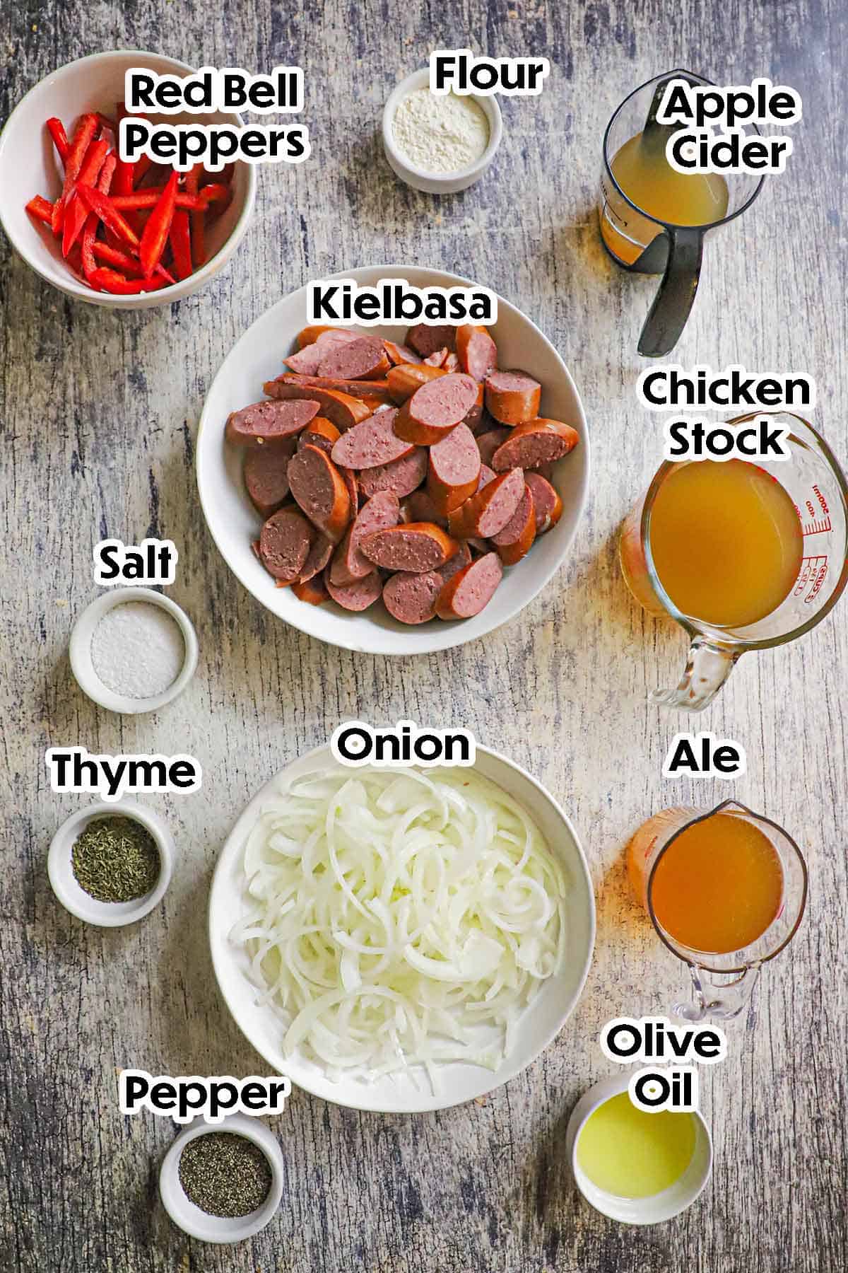 An arrangement of ingredients for beef-braised kielbasa with peppers on a grey wooden background including sliced kielbasa, sliced red bell peppers, chicken stock, apple cider, sliced onion, ale, flour, and seasonings.