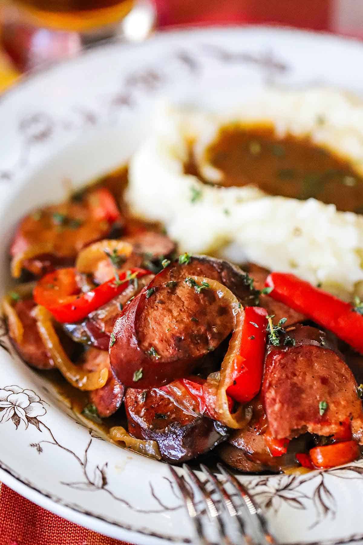 A close-up view of a vintage soup bowl filled with beer-braised kielbasa and peppers next to a pile of mashed potatoes with gravy on top.