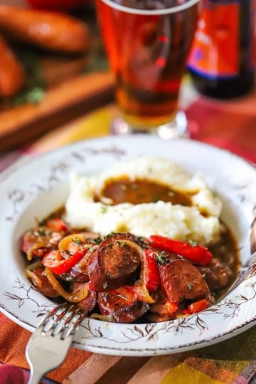 A vintage soup bowl filled with a serving of beer-braised kielbasa and peppers with a pile of mashed potatoes next to it in the bowl.