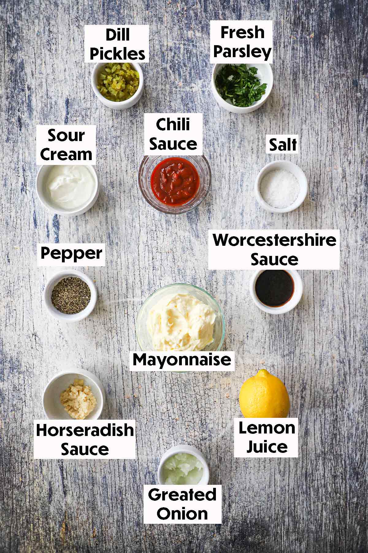An arrangement of ingredients for homemade Russian dressing on a wooden grey background including mayonnaise, chili sauce, sour cream, Worcestershire sauce, horseradish sauce, onion, lemon, pickles, parsley, salt, and pepper.