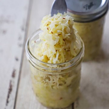 A person using a fork to hold up a bunch of homemade sauerkraut from a glass jar with another jar of sauerkraut in the background.
