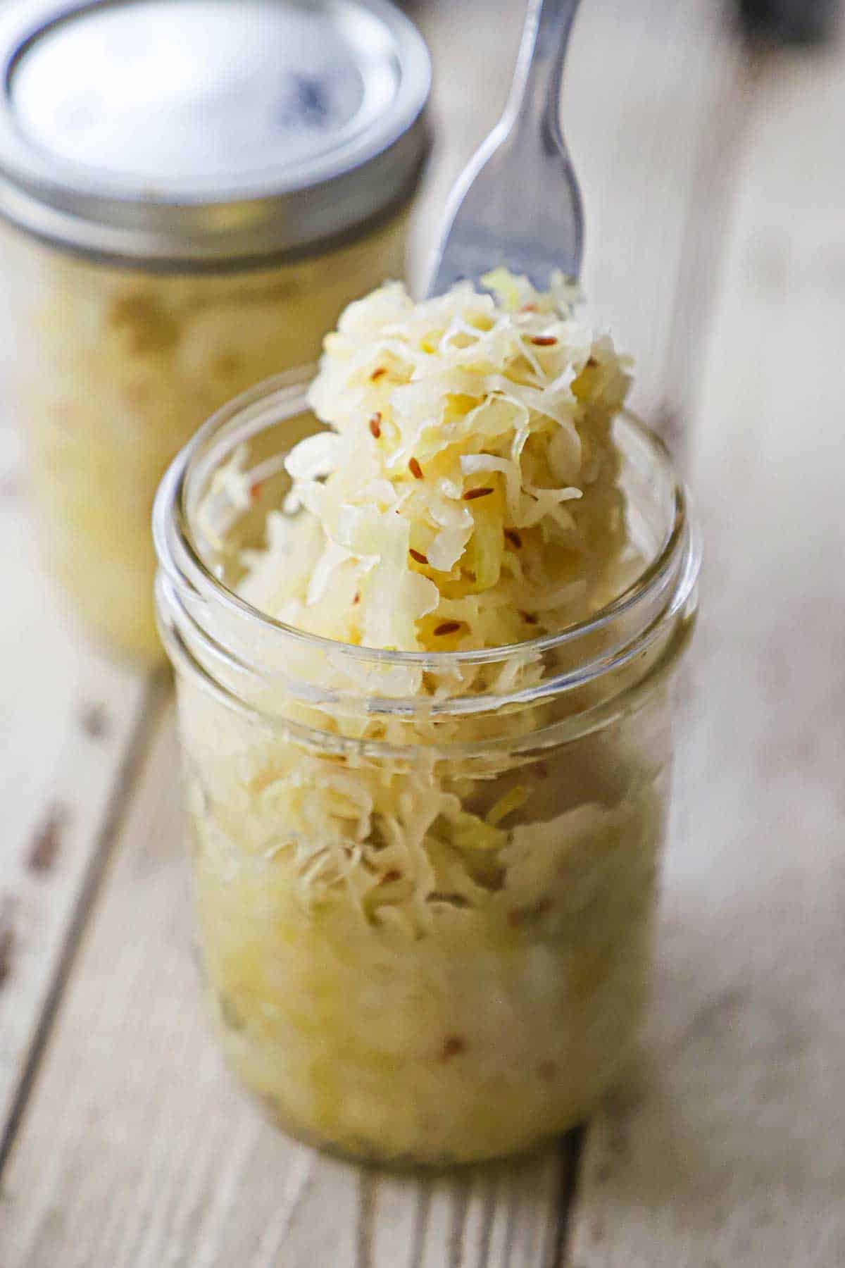 A person using a fork to hold up a bunch of homemade sauerkraut from a glass jar with another jar of sauerkraut in the background.