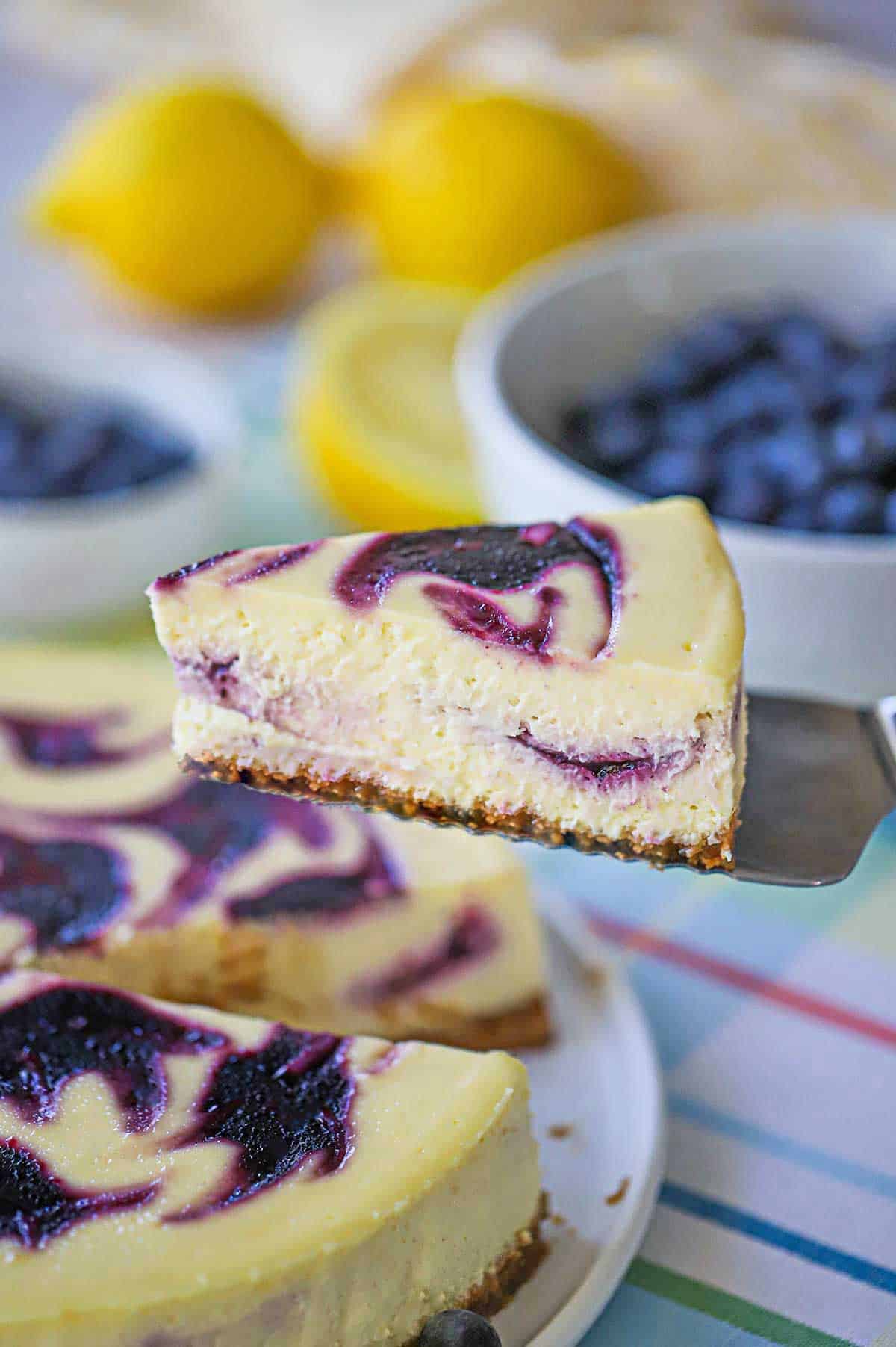 A person using a cake spatula to lift a slice of lemon blueberry cheesecake from a the cake resting on a circular white cake dish.