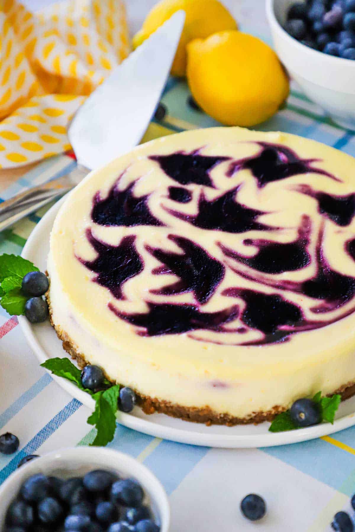 A lemon blueberry cheesecake with colorful blueberry-colored swirl designs on the top of the cake sitting next to a bowl of blueberries and two whole lemons.
